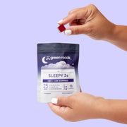 Hands holding a Green Roads Sleepy Z’s 10-count CBD + CBN gummies pouch and a dark red gummy against a lavender background.