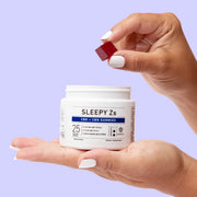 Hands holding an open Green Roads Sleepy Z’s CBD + CBN gummies jar and a blackberry-colored gummy on a lavender background.