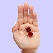 Open palm holding two dark red, cube-shaped Green Roads Sleepy Z’s gummies against a lavender background.