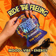 Hands opening a Ferris Wheel Sour Tropical gummies pouch on an orange beach towel beside sunglasses.
