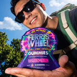 Person smiling in sunglasses and a backpack, holding a Ferris Wheel Watermelon 10-count gummies pouch outdoors