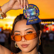 Person smiling and holding a Ferris Wheel Juicy Apple two-tablet pouch over one eye, with palm trees behind them.