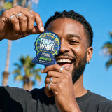 Person smiling and holding a Ferris Wheel Juicy Apple two-tablet pouch over one eye, with palm trees behind them.