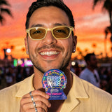 Person smiling in yellow sunglasses, holding a Ferris Wheel Pink Stardust two-tablet pouch at an outdoor event at sunset.