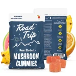 Road Trip Desert Stardust Mushroom Gummies pouch in Sunset Punch flavor, with front and back of the blue bag shown, surrounded by images of watermelon, orange slices, and pineapple pieces. Orange sugar-coated gummies placed in front of the pouch.